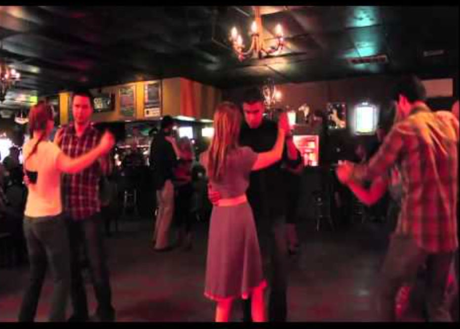 Couples dancing at a social dance night in a dimly lit bar-style dance hall with chandeliers and red mood lighting.