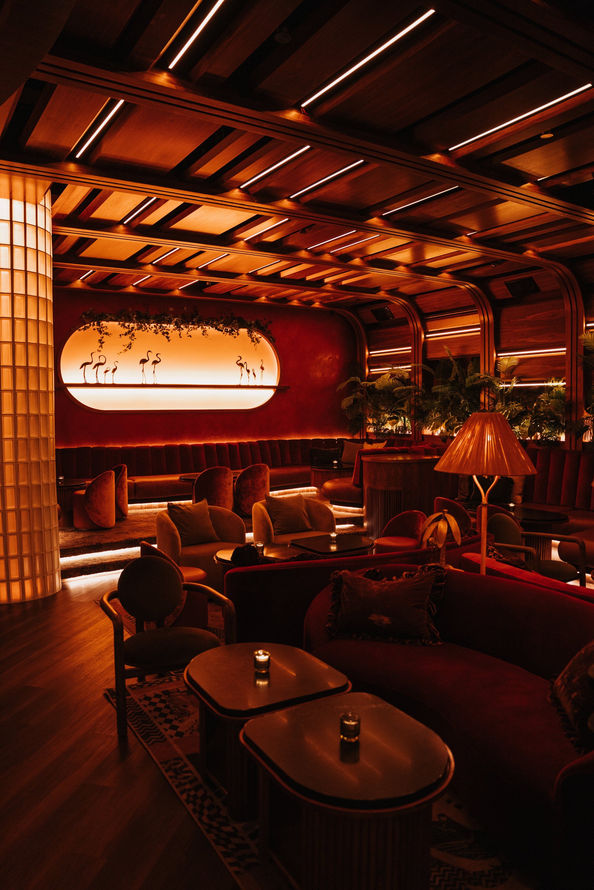Cozy upscale cocktail lounge interior with mid-century wood ceiling, plush velvet sofas and curved booths, moody amber lighting, candlelit tables, and silhouetted flamingo wall art.