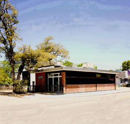 Small-town corner brick commercial building with flat roof and dark windows, shaded by oak trees under a clear blue sky