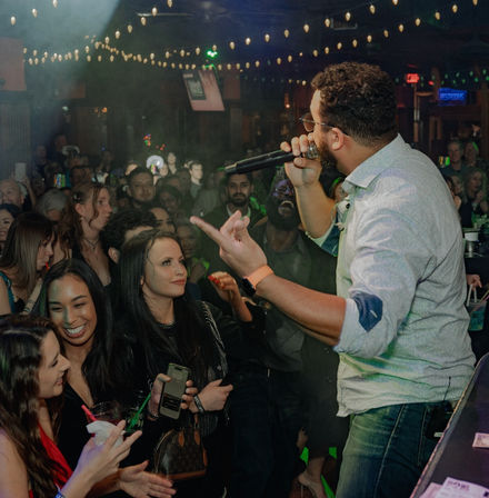 Live singer performing at a packed downtown nightclub, holding a microphone and engaging a cheering crowd under string lights as smiling patrons near the stage hold drinks and cheer.