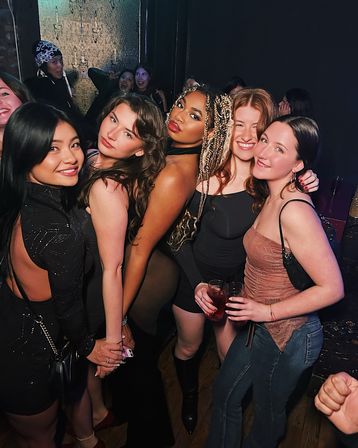 Five women posing closely in a lively nightclub group photo, dressed in party outfits with braided and loose hairstyles, smiling and holding drinks on a crowded dance floor during a girls' night out.