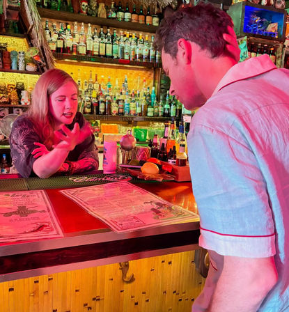 Bartender animatedly explaining the cocktail menu to a customer at a neon-lit tiki bar counter with rows of bottles and fresh garnishes.