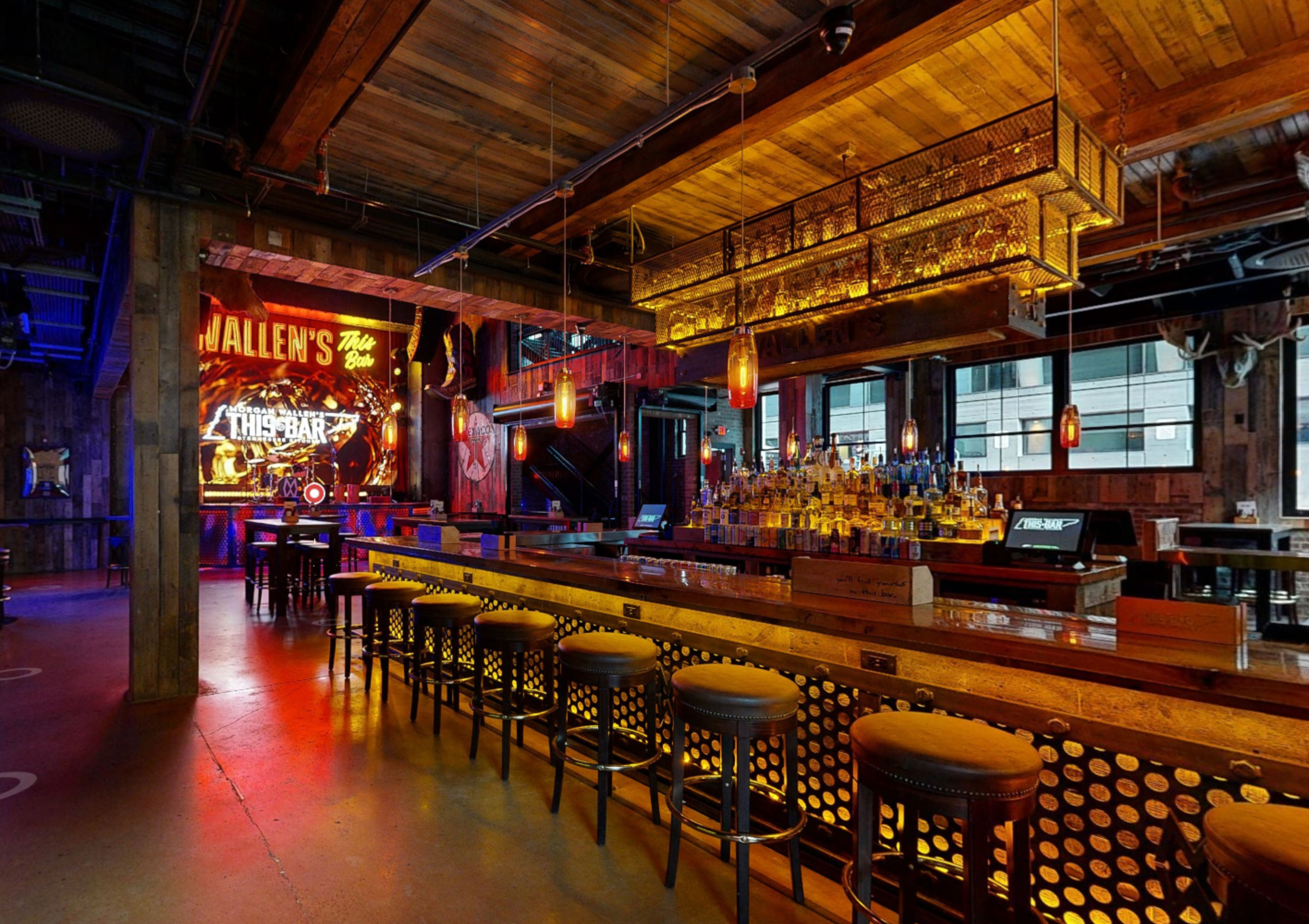 Industrial-style urban bar interior with a long wooden counter and metal stools, backlit bottle shelves, amber pendant lights, exposed wooden beams, stage with neon signage and large windows — cozy downtown nightlife vibe.