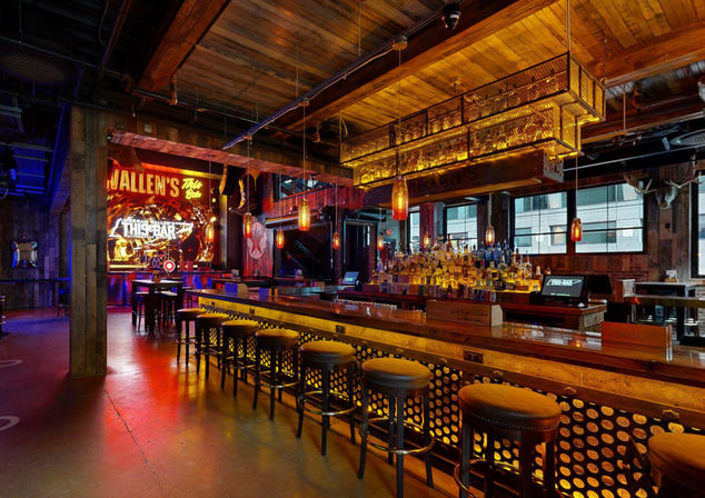 Industrial-style urban bar interior with a long wooden counter and metal stools, backlit bottle shelves, amber pendant lights, exposed wooden beams, stage with neon signage and large windows — cozy downtown nightlife vibe.
