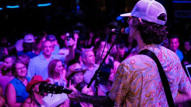 Singer with guitar in a baseball cap performing into a microphone on a purple-lit stage, packed crowd of concertgoers cheering and dancing close to the stage — lively live music nightlife scene.
