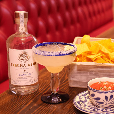 Salt-rimmed margarita with lime on a wooden table beside a tequila bottle, basket of tortilla chips and bowl of salsa in a cozy Mexican restaurant booth