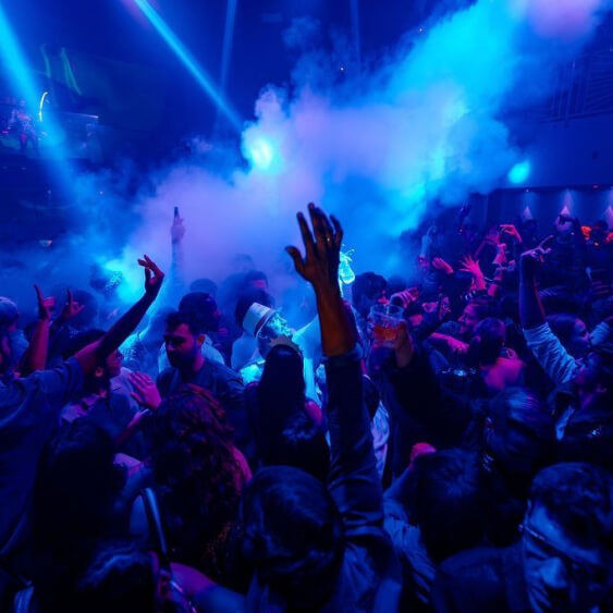 Packed city nightclub dance floor at night, energetic crowd with raised hands, blue laser lights and swirling fog from a fog machine