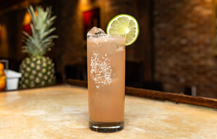 Tall pink citrus cocktail in a highball glass with a salt rim and lime wheel garnish, sitting on a marble bar counter with a pineapple and warm brick wall bokeh in the background.