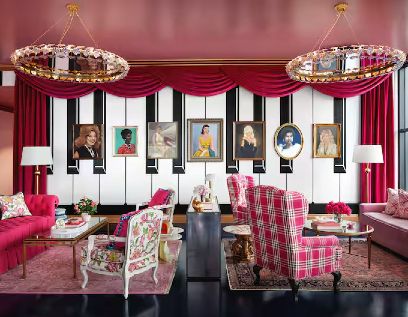 Playful pink luxury living room interior with red velvet swags, piano-key gallery wall of portraits, crystal ring chandeliers, pink velvet sofa, plaid armchair and floral accent chairs.