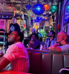 Tiki-themed bar interior with colorful hanging lanterns and neon lights, a group of patrons at a booth watching a screen and sipping cocktails in a lively nightlife setting.