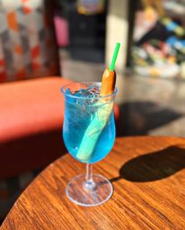 Sunlit bright-blue cocktail with ice in a stemmed glass on a wooden café table, garnished with a small sausage skewered by a green straw.