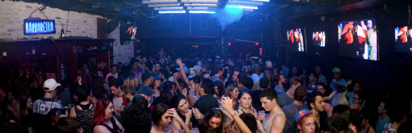Packed indoor nightclub dance floor under blue neon lights, people dancing and holding drinks with video screens along the wall.