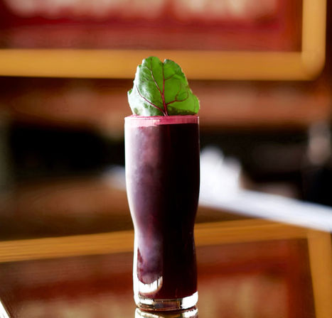 Vibrant dark purple beetroot cocktail in a tall glass topped with pink foam and a green leaf garnish, sitting on a polished wooden bar table