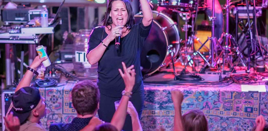 Female singer passionately performing on a colorful-lit stage with drums and band gear behind her, while an enthusiastic crowd reaches up and cheers at a lively local live music venue