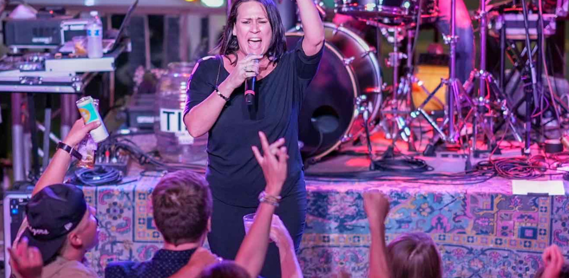 Female singer passionately performing on a colorful-lit stage with drums and band gear behind her, while an enthusiastic crowd reaches up and cheers at a lively local live music venue