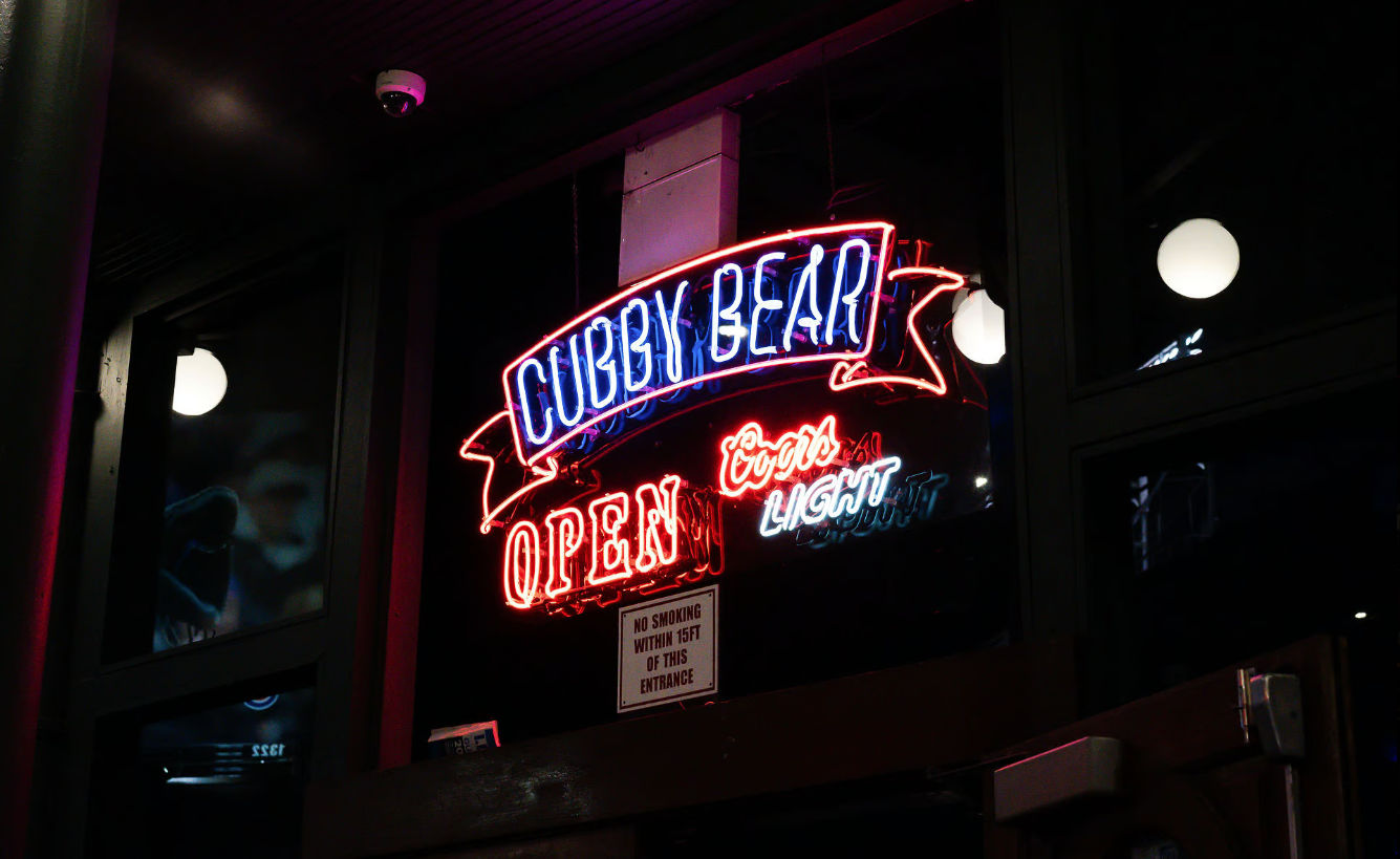 Glowing neon bar sign in blue and red displaying OPEN above a dark storefront entrance, reflected in windows with round globe lights at night