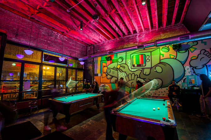 Neon-lit downtown pool hall with two billiard tables, exposed brick and wooden-beam ceiling, a large colorful cartoon TV-head mural and blurred patrons playing and socializing
