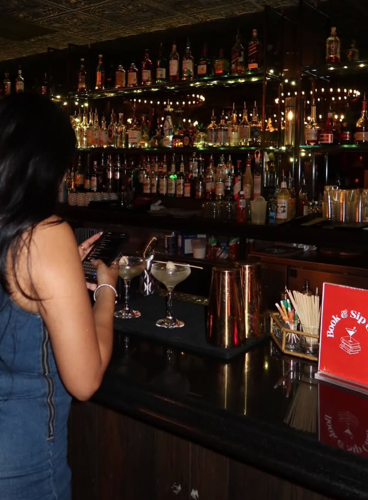 Patron at a dimly lit cocktail bar tapping a phone at a polished black counter with two martini glasses, copper shakers and mirrored liquor shelves strung with lights