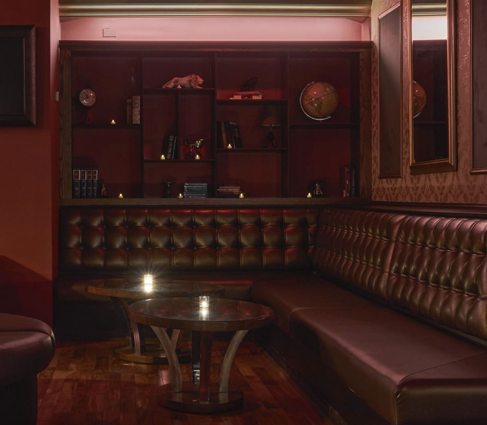Moody, intimate cocktail lounge with tufted brown leather banquette, low wooden tables lit by candles and shelves with books and a globe.