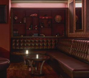 Moody, intimate cocktail lounge with tufted brown leather banquette, low wooden tables lit by candles and shelves with books and a globe.
