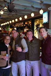 Four young men smiling and posing at a lively indoor bar/arcade with hanging globe lights, festive holiday lights, and drinks.