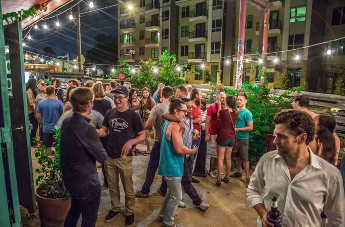 Lively outdoor urban patio at night with string lights and a crowd of young adults socializing and drinking in front of apartment buildings