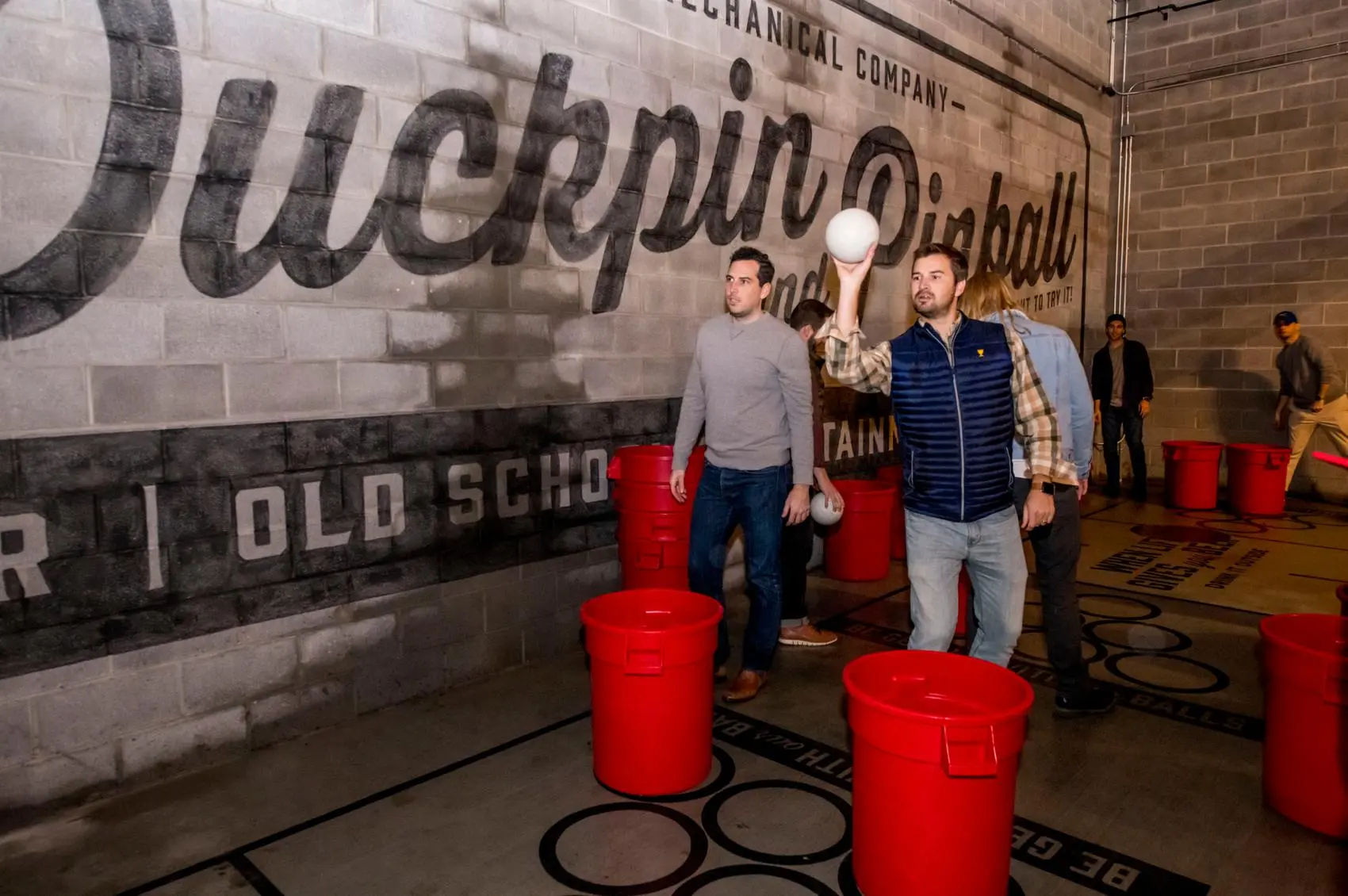 Group of casually dressed men tossing white balls into red plastic bins in an indoor recreational space with concrete block walls and a large painted mural.