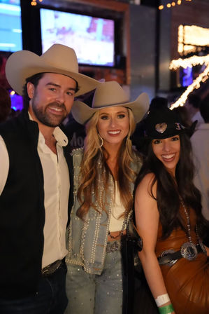 Three smiling people in cowboy hats at a lively country bar — studded denim jacket, leather outfit, string lights and TV in the background