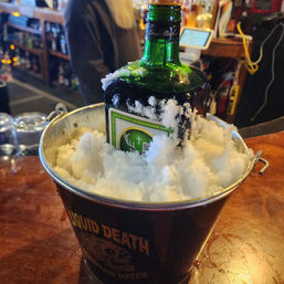 Green liqueur bottle chilled upright in a black metal bucket filled with crushed ice on a wooden bar counter in a busy pub setting