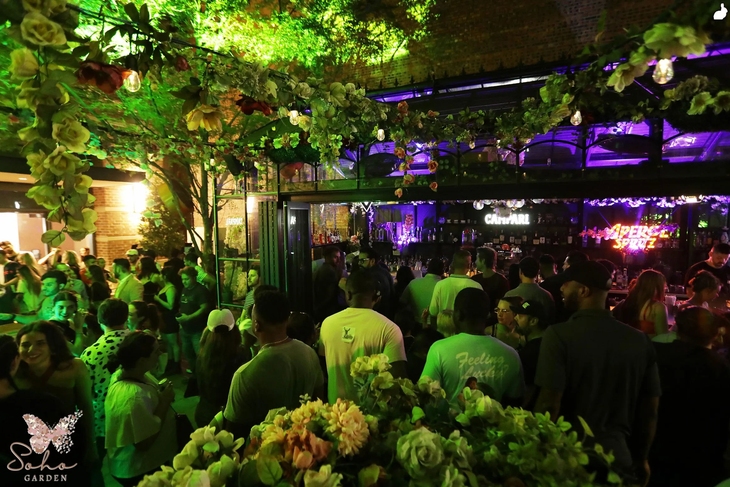 Crowded outdoor garden bar at night under a flower-covered canopy, green and purple lighting, a neon drink sign, and patrons gathered around the illuminated bar.
