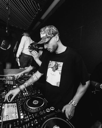 Black-and-white photo of two DJs in a nightclub booth — one wearing a camo cap with a tattooed arm adjusting knobs on turntables and digital controllers while a partner listens on headphones.