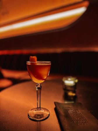 Amber cocktail in a coupe glass with an orange peel garnish on a dimly lit bar table, warm orange lounge lighting and candle glow creating a cozy nightlife vibe.