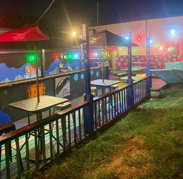 Nighttime outdoor patio with umbrella-covered tables, multicolored string lights, painted mural and wooden railing overlooking a grassy lawn.