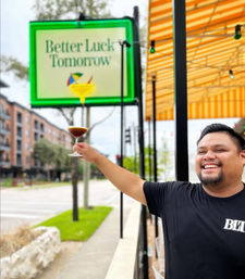 Smiling man toasting with a martini glass on a sunny sidewalk patio beneath a yellow-striped awning, green-framed sign and urban street in the background.