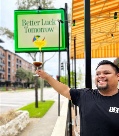 Smiling man toasting with a martini glass on a sunny sidewalk patio beneath a yellow-striped awning, green-framed sign and urban street in the background.