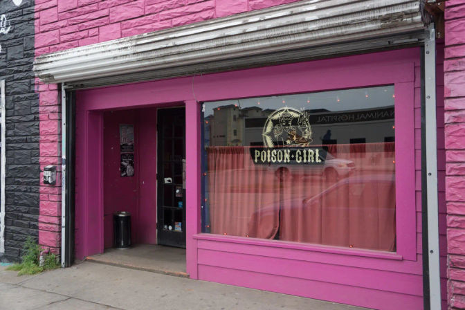 Bright magenta storefront on an urban sidewalk featuring a retro circular window decal, coral curtains and an open doorway.