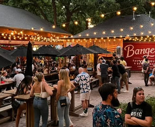 Lively outdoor beer garden patio at dusk with string lights, picnic tables and umbrellas, people socializing and enjoying food and drinks.