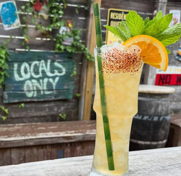 Tall tropical cocktail with crushed ice, green bamboo straw, chili-salt rim, orange slice and fresh mint, on a weathered wooden outdoor bar with a rustic fence, ivy and a painted "LOCALS ONLY" sign in the background.