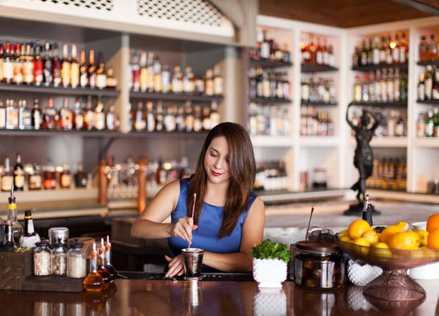 Mixologist stirring a cocktail at a polished bar counter with stocked liquor shelves, jars of garnishes, fresh lemons and a small herb pot.
