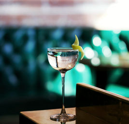 Cocktail coupe glass with clear drink and a zesty lemon twist perched on a polished bar counter, teal tufted banquette and soft bokeh lights in the background