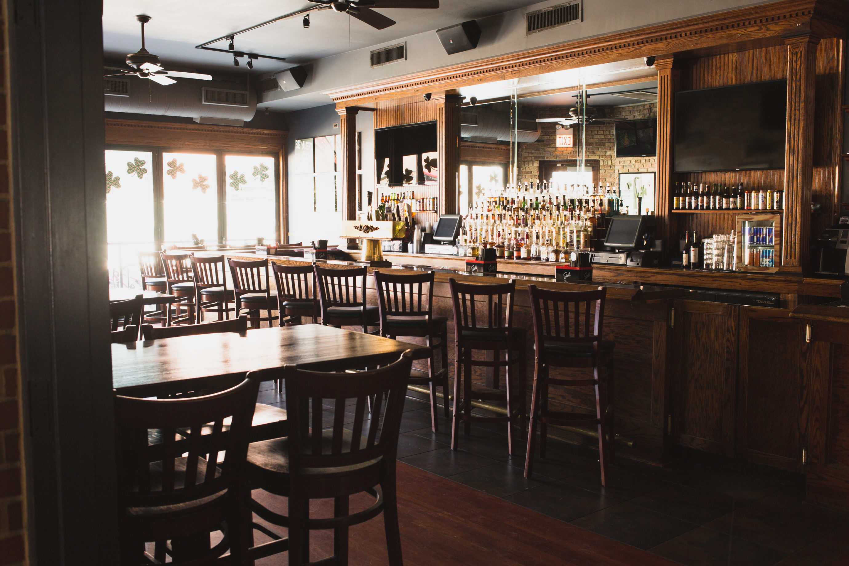 Sunlit cozy pub interior with a long wooden bar, row of high stools, mirrored shelves stocked with liquor bottles, and wooden tables and chairs.