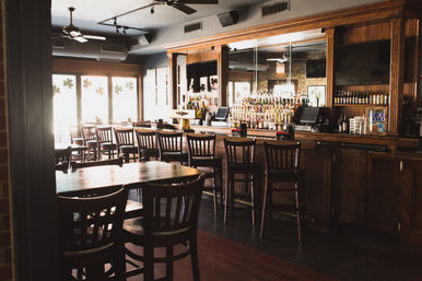 Sunlit cozy pub interior with a long wooden bar, row of high stools, mirrored shelves stocked with liquor bottles, and wooden tables and chairs.