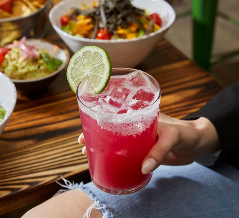 Bright pink margarita-style cocktail with ice, salted rim and lime wheel garnish, held over a wooden restaurant table with bowls of chips and salad blurred in the background