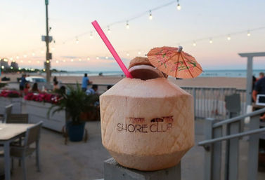 Tropical coconut drink with pink straw and paper umbrella on a beachside deck at sunset, string lights and ocean in the background