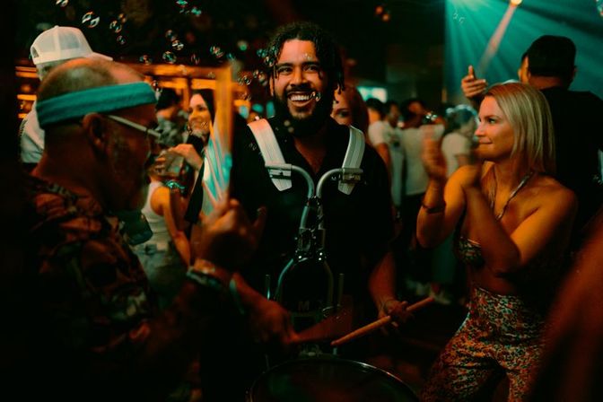 Smiling drummer in a harness plays a marching drum while partygoers clap and bubbles float on a crowded nightclub dance floor under colorful lights.