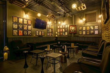 Cozy industrial-style lounge interior with leather sofas and armchairs around small tables, exposed ceiling and warm hanging bulbs, brick wall and gallery wall of framed vintage posters and a wall-mounted TV.