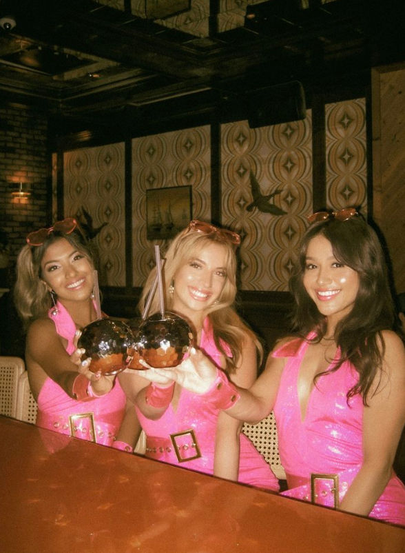Three smiling women in matching hot-pink dresses toasting with mirrored disco-ball cocktail mugs at a retro-styled bar with patterned wallpaper — girls' night out.
