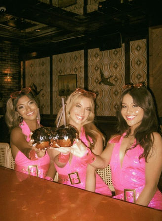 Three smiling women in matching hot-pink dresses toasting with mirrored disco-ball cocktail mugs at a retro-styled bar with patterned wallpaper — girls' night out.