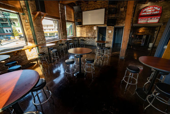 Cozy downtown brick-walled sports bar interior with polished dark floors, round high-top tables and metal stools, large street-facing windows and a neon sports sign.