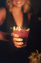Hand holding a pink cocktail in a rocks glass garnished with gummy candies on a skewer, dimly lit bar/nightlife scene.
